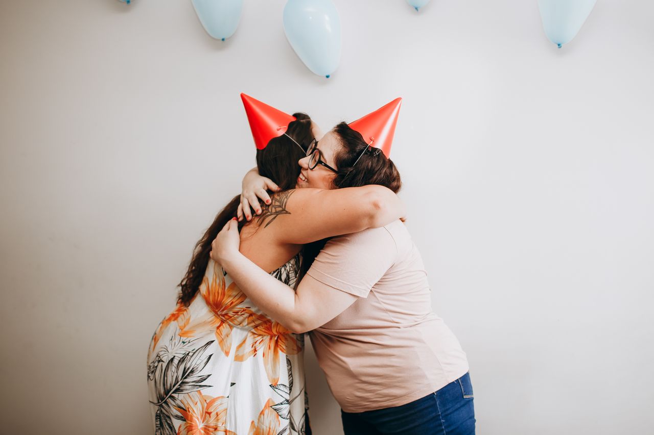 about-img Two women hugging at a birthday party with balloons and hats, capturing joy and togetherness.