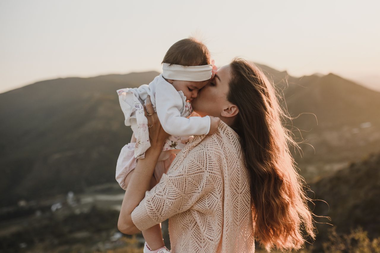 ours-journey A mother gently kissing her baby while standing outdoors with a mountainous backdrop.